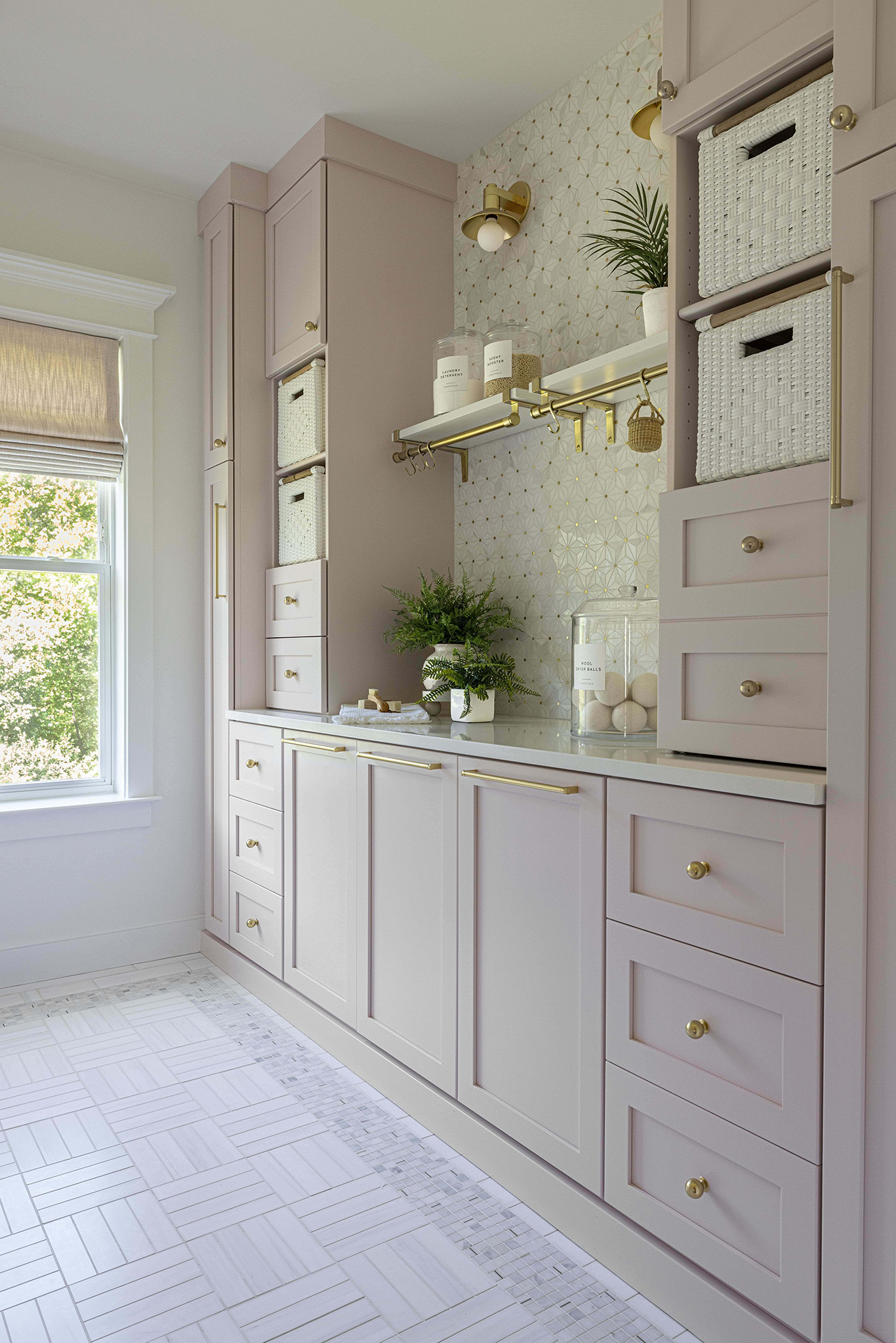 Luxe laundry room custom cabinetry in soft blush taupe with woven storage baskets, brass rail hanging rack, and marble mosaic wall tile by Sho and Co Cabinetry